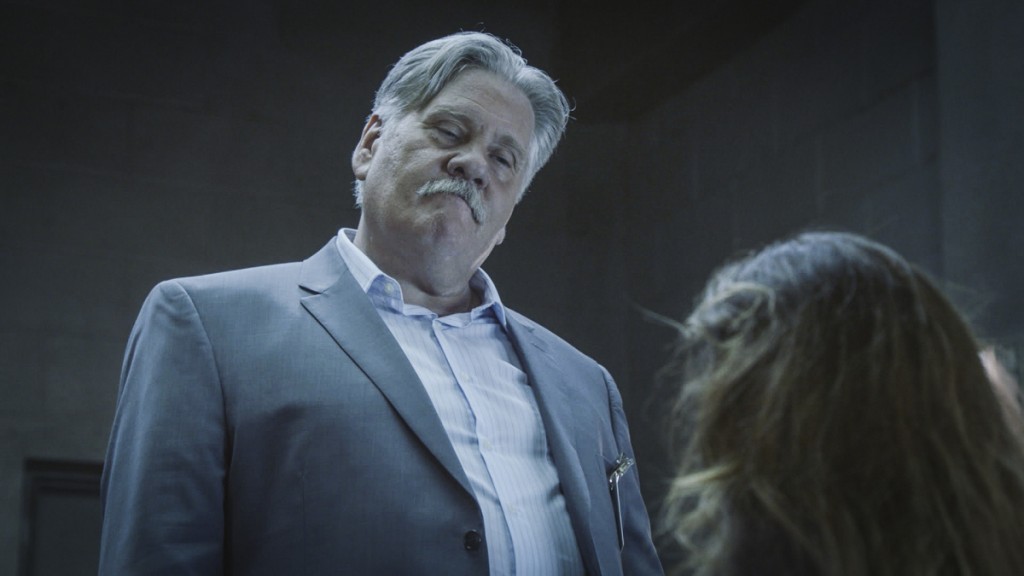 Harry (William Forsythe) pose des questions à une femme dans la salle d'interrogatoire.