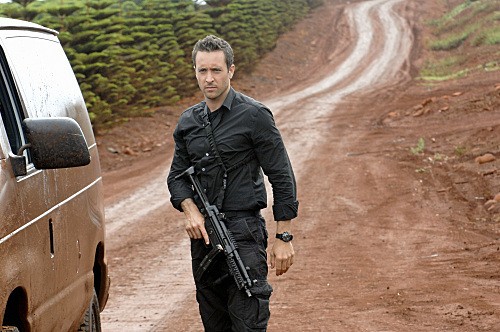 McGarrett se trouve au milieu d'un chemin en terre, à côté d'un van, avec une arme à la main.