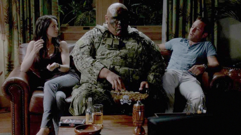 Catherine (Michelle Borth) et Steve (Alex O'Loughlin) fixent Kamekona (Taylor Wily) qui est assis au milieu du canapé avec un bol de pop-corn.