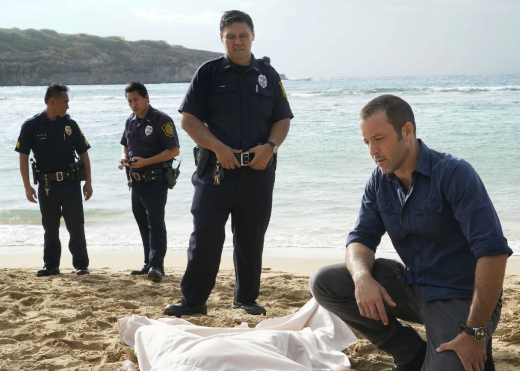 Sur une plage avec Pua Kai (Shawn Thomsen), Steve (Alex O'Loughlin) est agenouillé devant un cadavre recouvert d'un drap blanc.