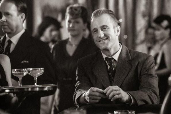 En 1941, Milton Cooper (Scott Caan) est installé à une table et contemple une personne en souriant.