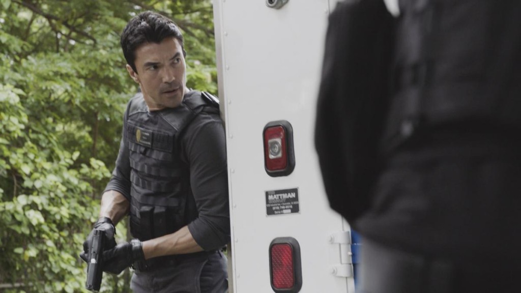 Adam Noshimuri (Ian Anthony Dale) se trouve sur le terrain. Il est armé et porte un gilet pare-balles.